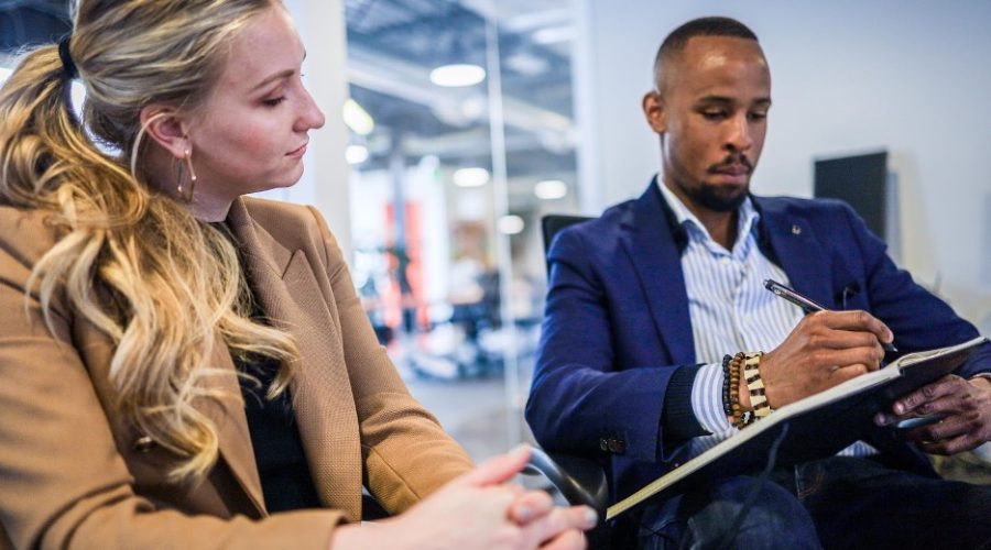 A white woman and an African American male are siting while the male is writing in a notebook