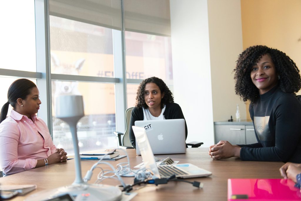 Three African American woman are sitting at desk talking. 