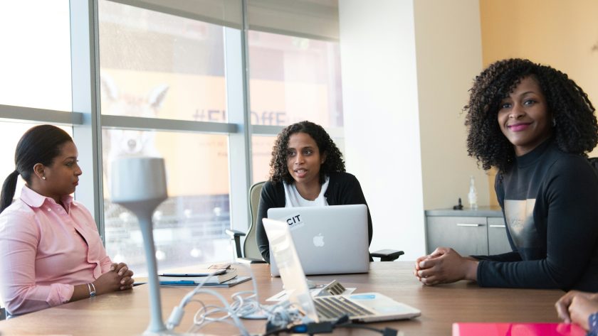 Three African American woman are sitting at desk talking.