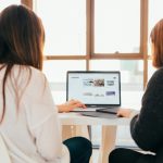 2 Woman are sitting at a desk looking at a computer screen