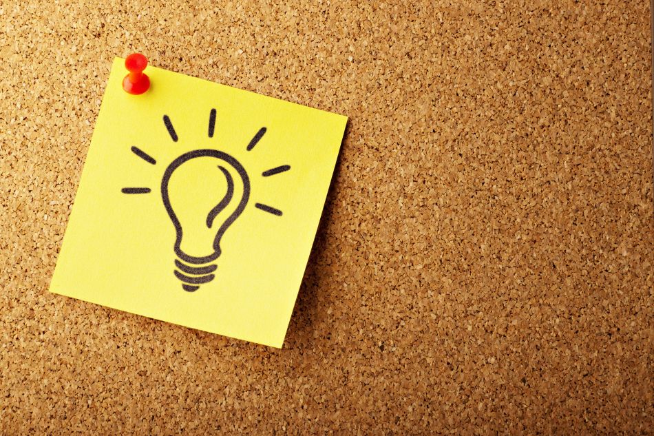 Yellow postit note with a lightbulb drawn on it, has a red thumbtack and is fastened to a cork board