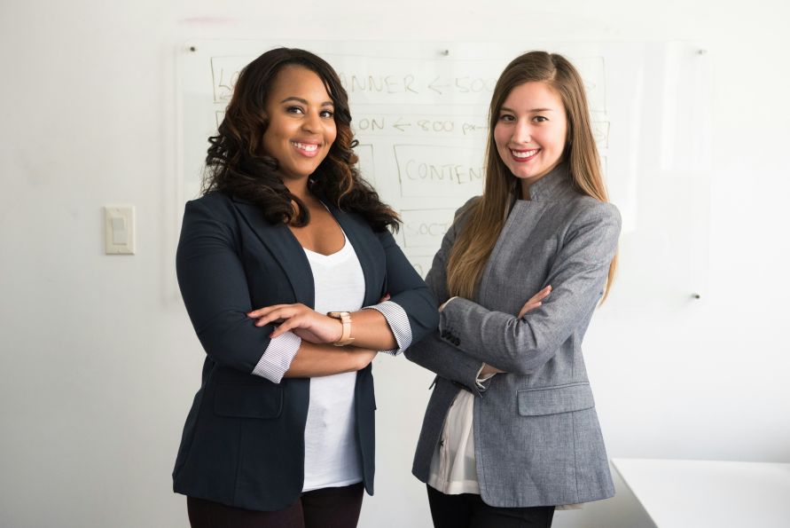 Two diverse women in dress clothes wmiling with arms crossed in front of white board